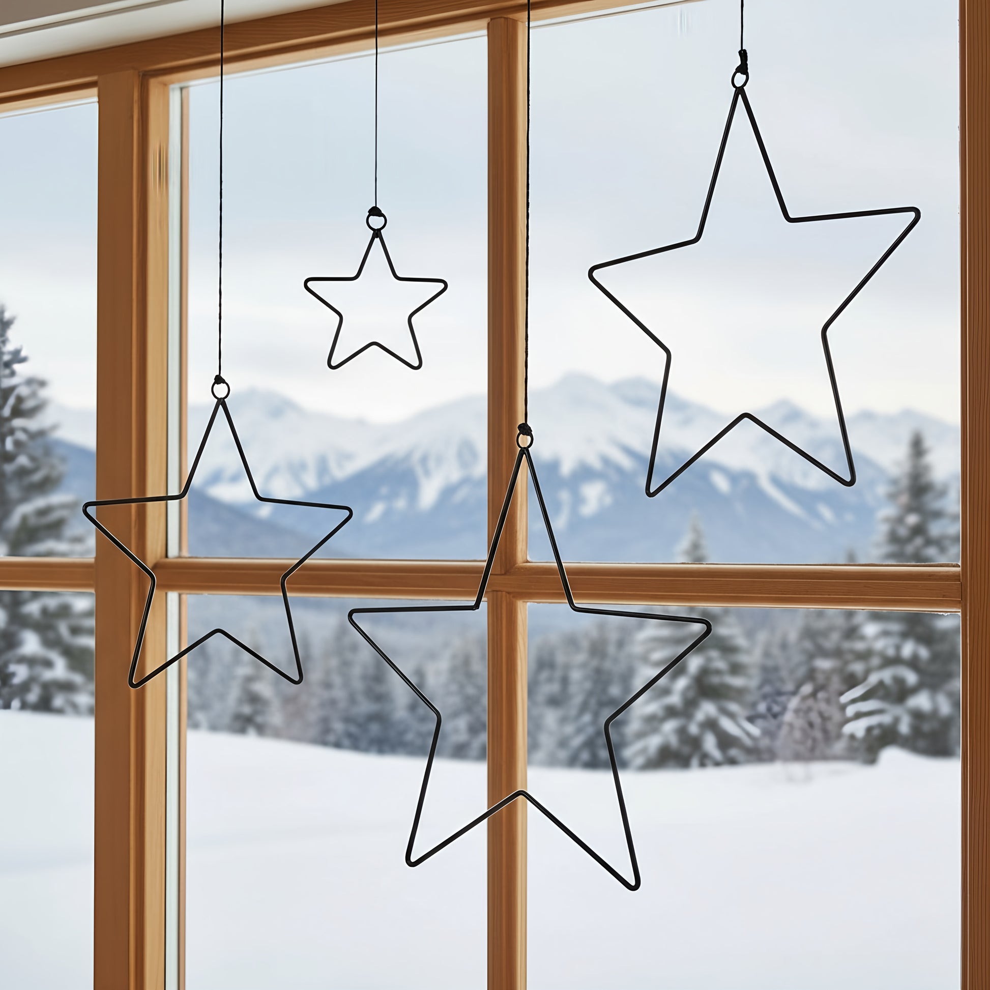 Vier schwarze Metallsterne in unterschiedlichen Größen hängen mit Schnur an einem Holzfenster. Draußen ist eine verschneite Winterlandschaft mit Tannenbäumen und Bergen im Hintergrund zu sehen. Die Sterne wirken schlicht und modern als weihnachtliche Fensterdekoration.