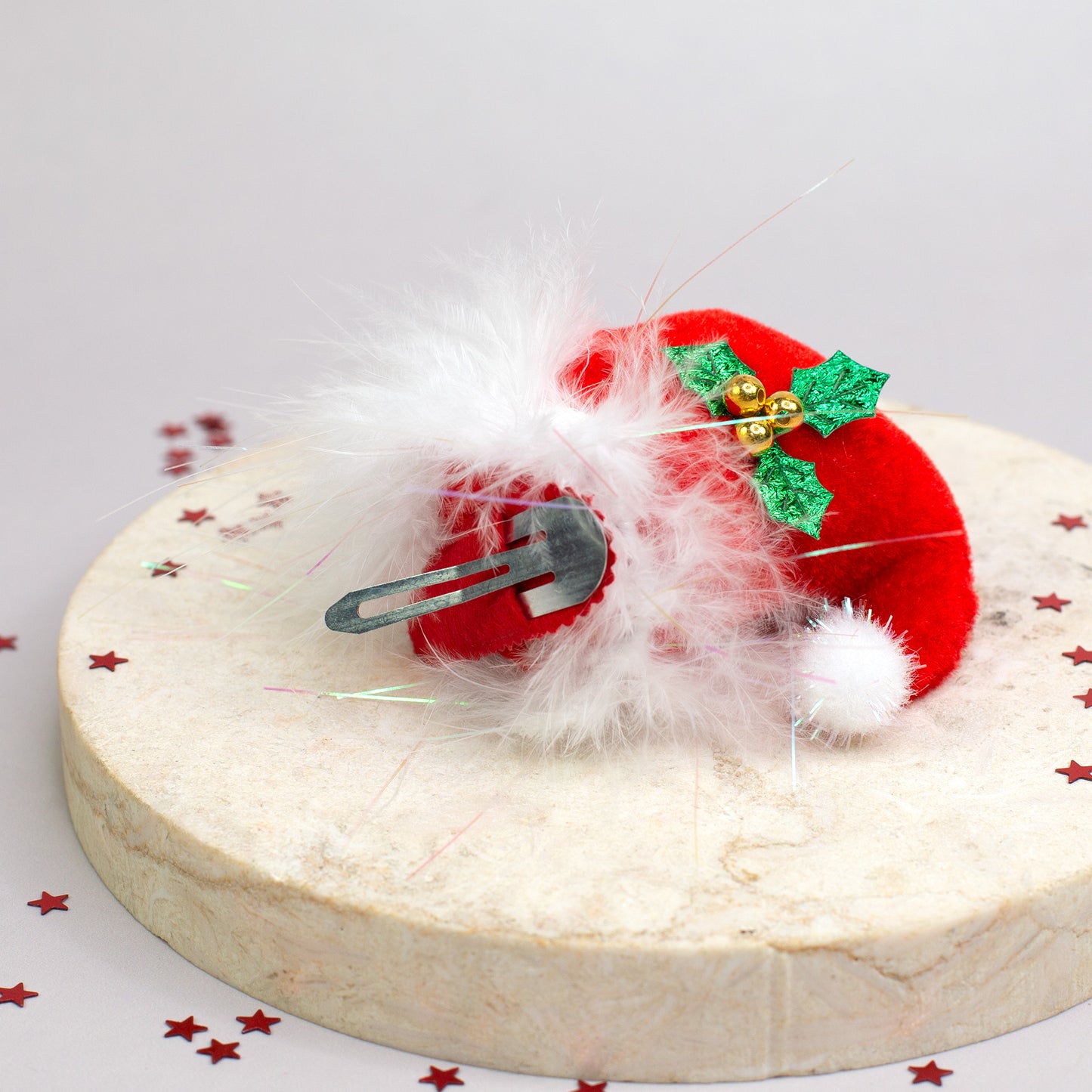 Roter Weihnachts-Haarclip aus Plüsch mit weißer Flauschkante, grünen Stechpalmenblättern und goldenen Glöckchen auf rundem Podest.