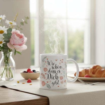 Dampfende Keramiktasse mit Spruch „Wie schön, dass es Dich gibt“ und Blumenmuster auf hell gedecktem Frühstückstisch.