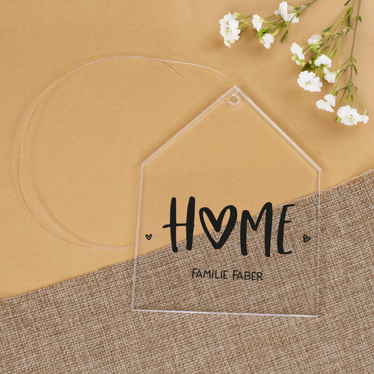 Transparenter Acrylanhänger in Hausform mit HOME-Aufdruck und personalisiertem Text Familie Faber auf Stoffunterlage mit weißen Blumen
