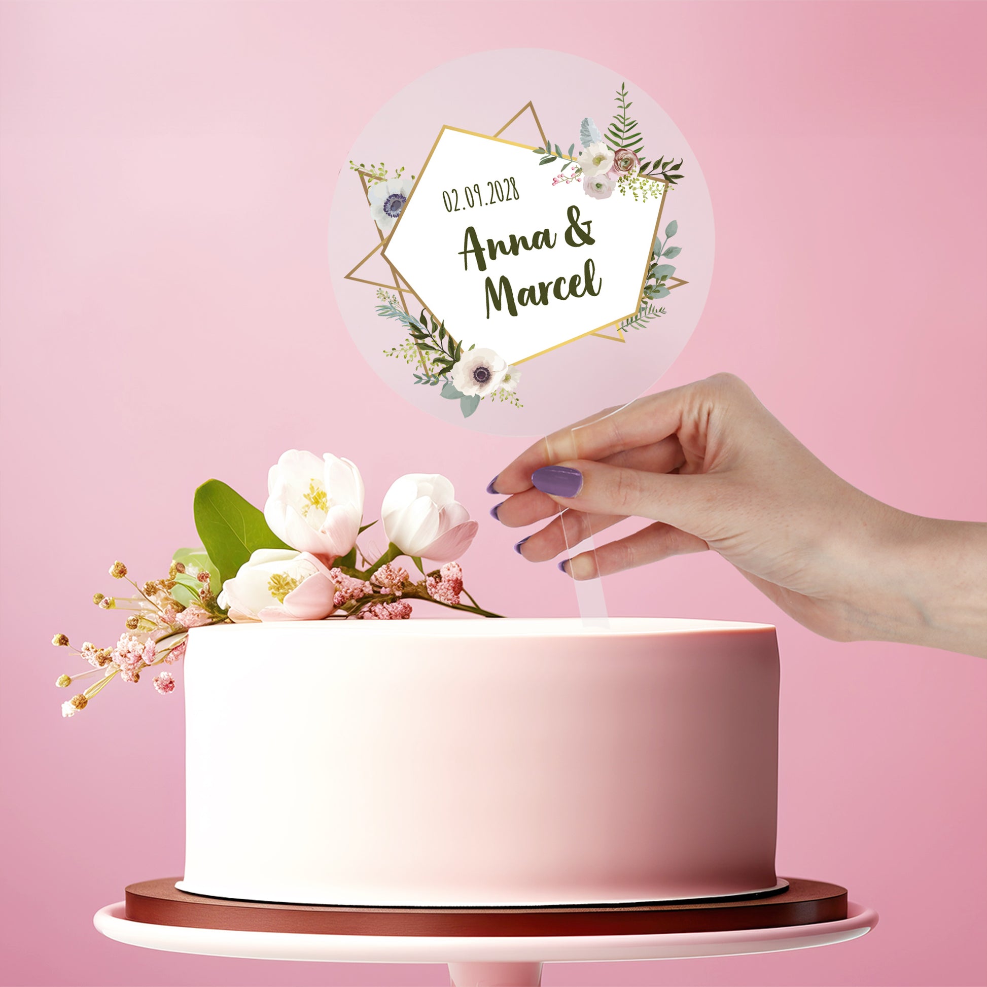 Personalisierter Tortentopper Hochzeit aus Acryl mit Namen Anna & Marcel und Datum auf weißer Torte vor rosa Hintergrund