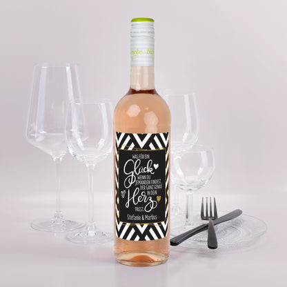 Roséweinflasche mit schwarz-weißem Etikett und goldener Schrift, Spruch „Was für ein Glück…“ und Namen Stefanie & Markus, vor Gläsern und Besteck dekoriert