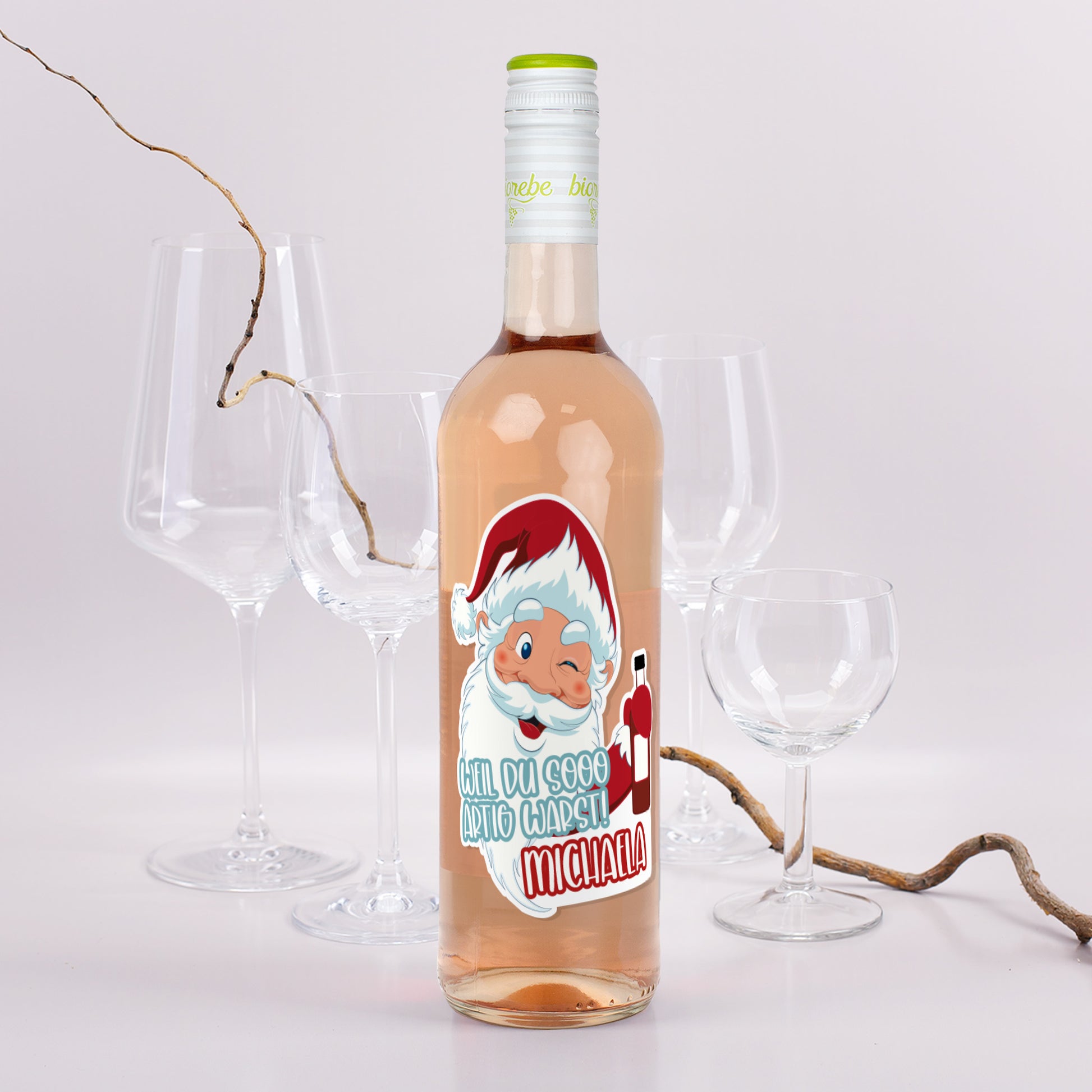 Roséweinflasche mit Weihnachtsmann-Etikett „Weil du sooo artig warst! Michaela“, dekoriert mit Gläsern und Zweigen.