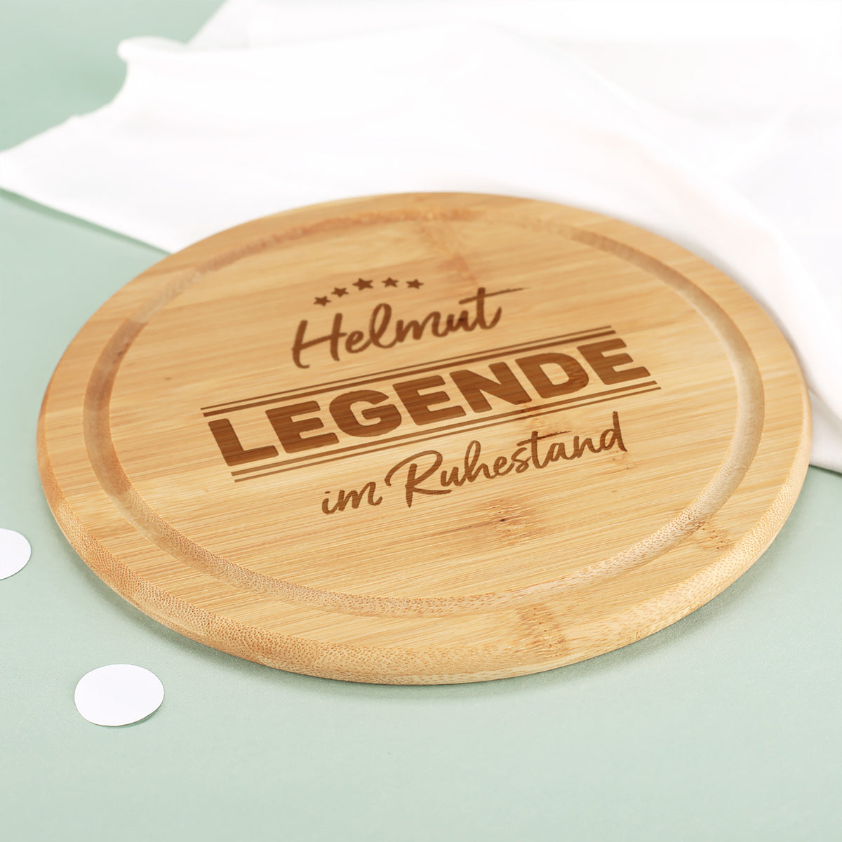 Schräg fotografiertes rundes Bambus Schneidebrett mit Gravur „Helmut – Legende im Ruhestand“ und sichtbarer Saftrille