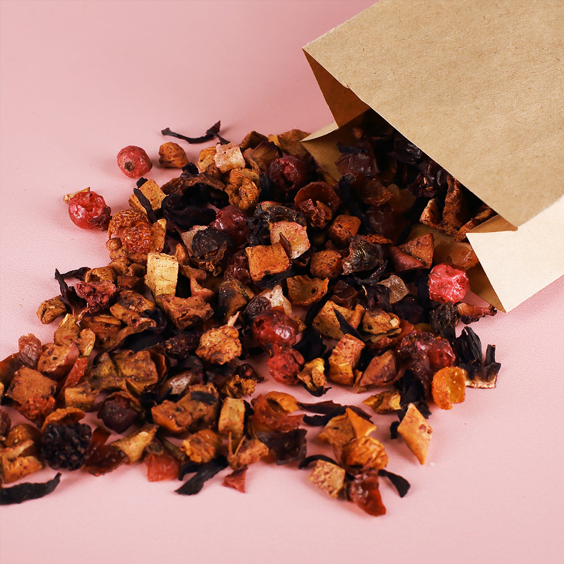 Aromatischer Früchtetee in loser Form, mit getrockneten Beeren und Fruchtstücken, aus einer braunen Kraftpapiertüte auf rosa Untergrund ausgeschüttet. Ideal für entspannende Teemomente.