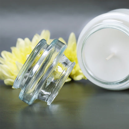 Duftkerze in einem transparenten Glas mit abnehmbarem Deckel, auf dunklem Untergrund mit gelben Blüten im Hintergrund. Perfekt für entspannende Momente und gemütliche Atmosphäre.