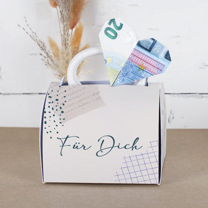 Fototasse in Geschenkverpackung mit „Für Dich“-Aufschrift, aus der ein Herz aus einem gefalteten 20-Euro-Schein ragt. Die Verpackung hat ein dezentes, modernes Design mit grafischen Elementen und wirkt wie eine kleine Handtasche.