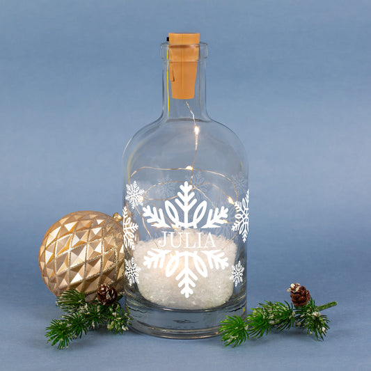 LED-Karaffe mit großer Schneeflocke, Kunstschnee und personalisiertem Namen vor blauem Hintergrund dekoriert mit Weihnachtskugel.