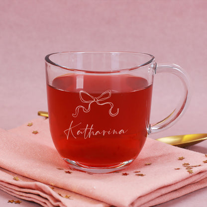 Gravierte Glastasse mit Schleifenmotiv und Name „Katharina“ auf rosa Hintergrund, gefüllt mit Tee