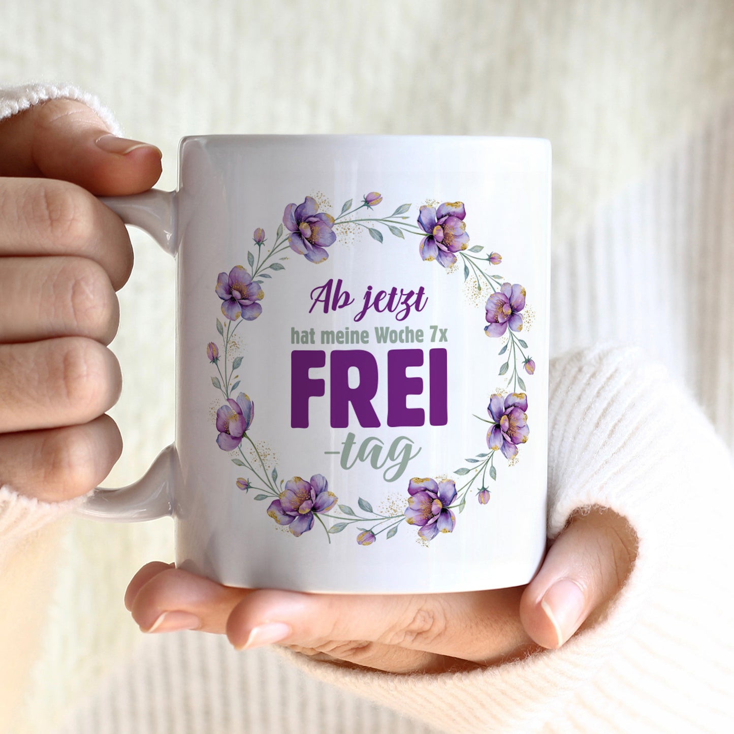Weiße Tasse mit Spruch „Ab jetzt hat meine Woche 7x Frei-tag“ und violettem Blumenkranz in den Händen gehalten