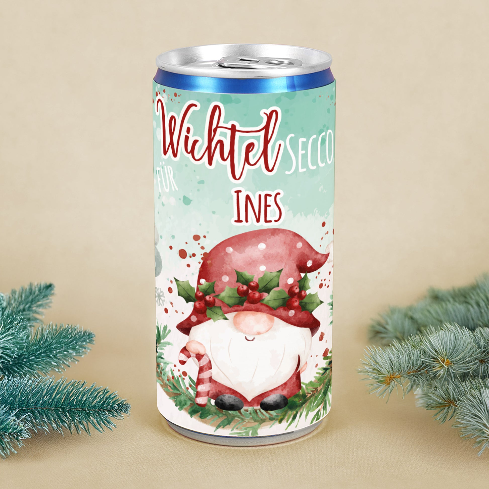 Personalisierte Secco-Dose mit Wichtelmotiv, roter Mütze, Mistelkranz und Name Ines, dekoriert mit Tannenzweigen als Weihnachtsgeschenk.