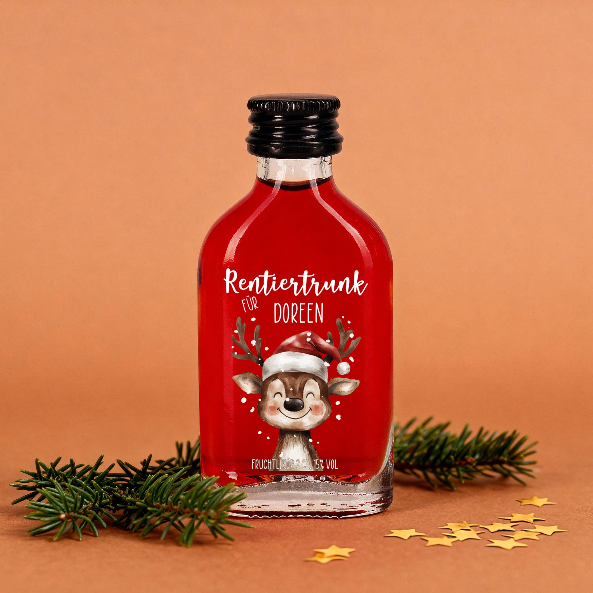 Kleine Glasflasche mit rotem Fruchtlikör, Aufdruck „Rentiertrunk für Doreen“ und fröhlichem Rentier mit Weihnachtsmütze, dekoriert mit Tannenzweigen und goldenen Sternen.