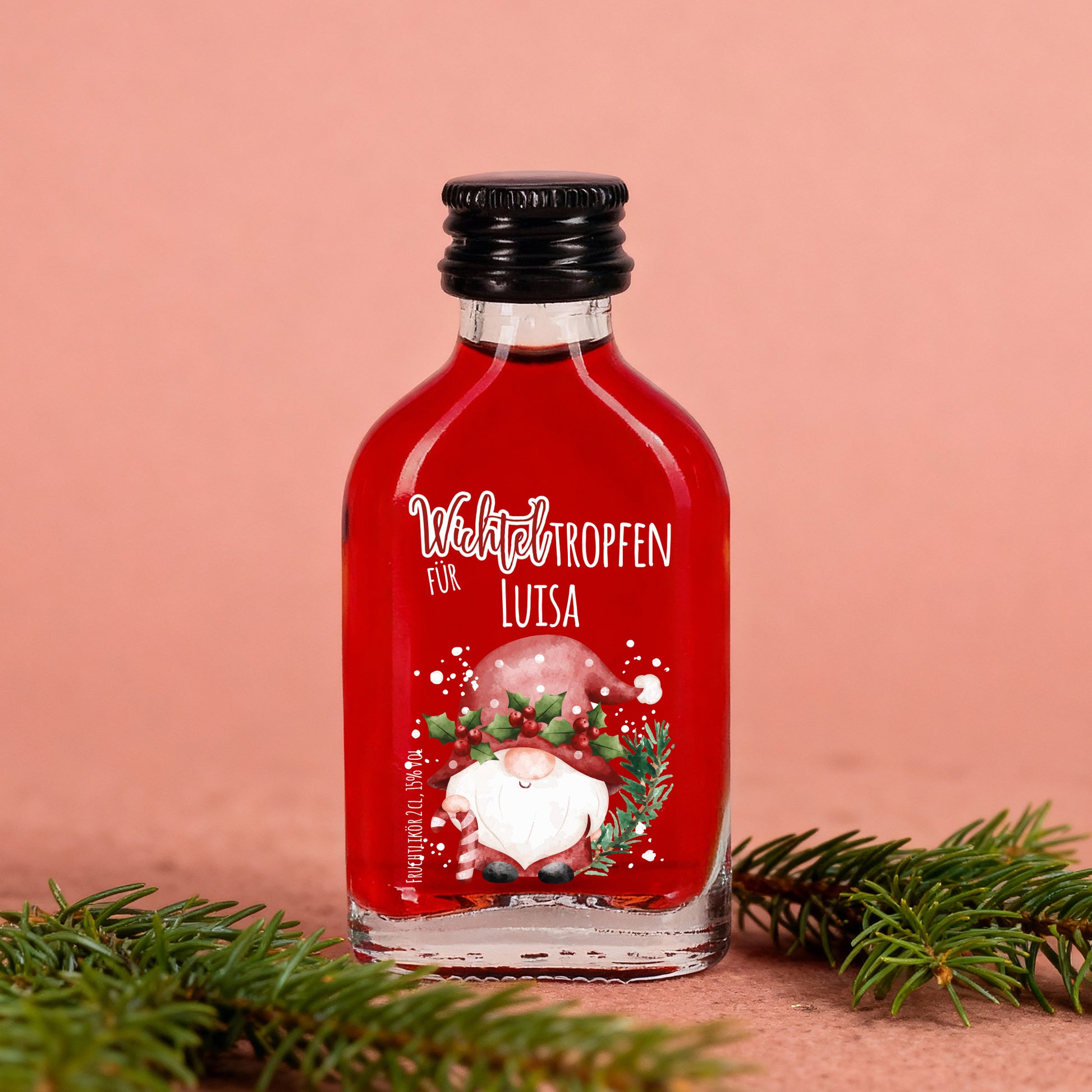 Wichteltropfen-Flasche mit rotem Likör und Namenspersonalisierung vor rosafarbenem Hintergrund, weihnachtlich dekoriert.