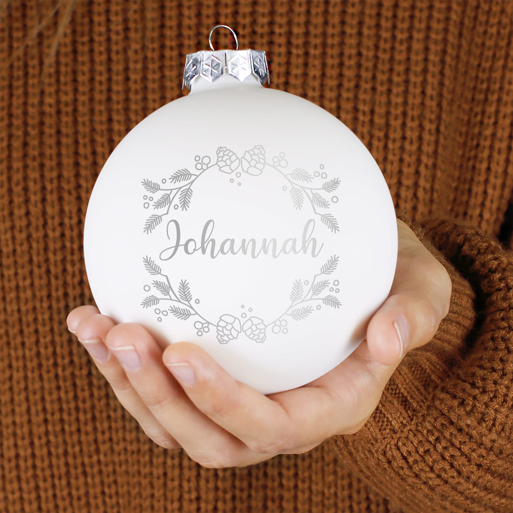 Weiße matte Weihnachtskugel mit personalisierter Namensgravur „Johannah“, verziert mit feinem Tannenzweigkranz und Zapfenmotiv, in zwei Händen gehalten.