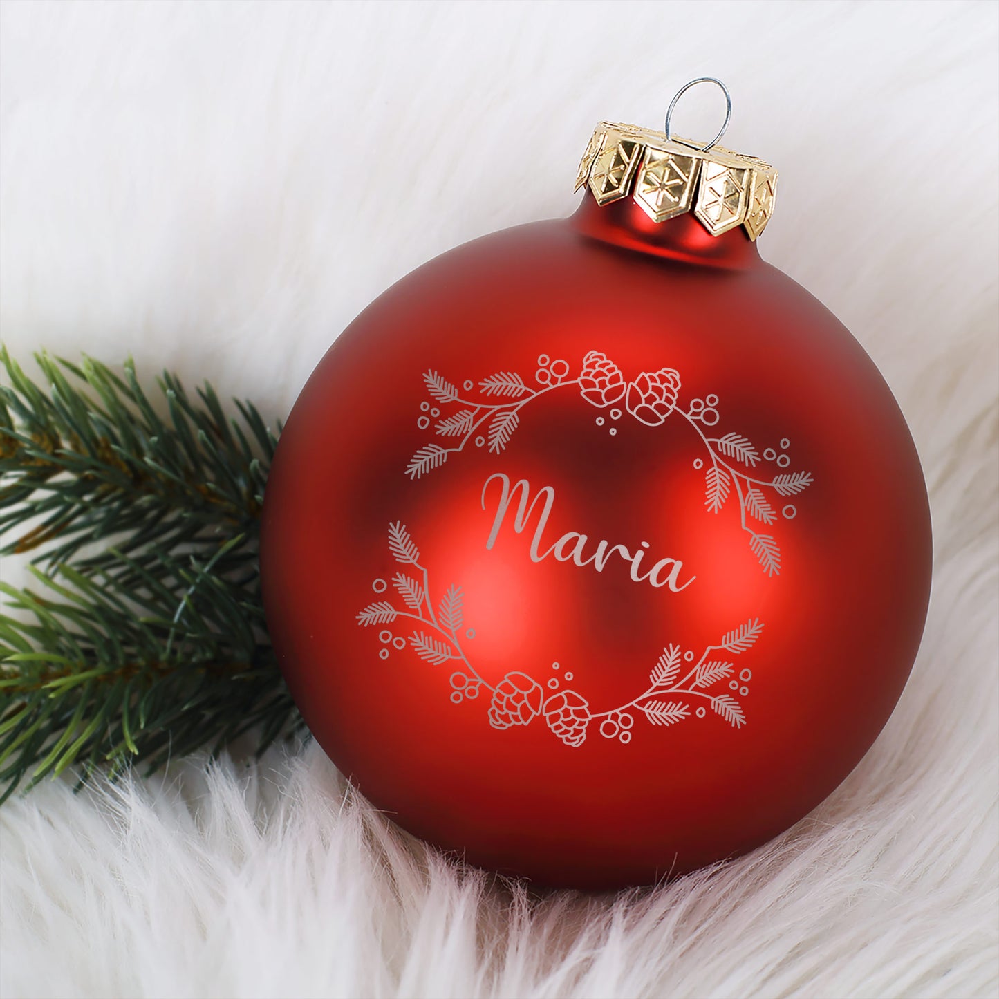 Rote matte Weihnachtskugel mit Gravur „Maria“ und filigranem Weihnachtskranzmotiv aus Tannenzweigen und Beeren, liegend auf weißem Fell mit Tannenzweig-Deko.