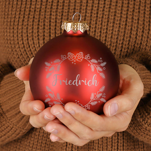 Rote Weihnachtskugel mit personalisierter Namensgravur „Friedrich“, verziert mit filigranem Tannenzweig-Kranzmotiv, glänzende Oberfläche, in Händen gehalten.