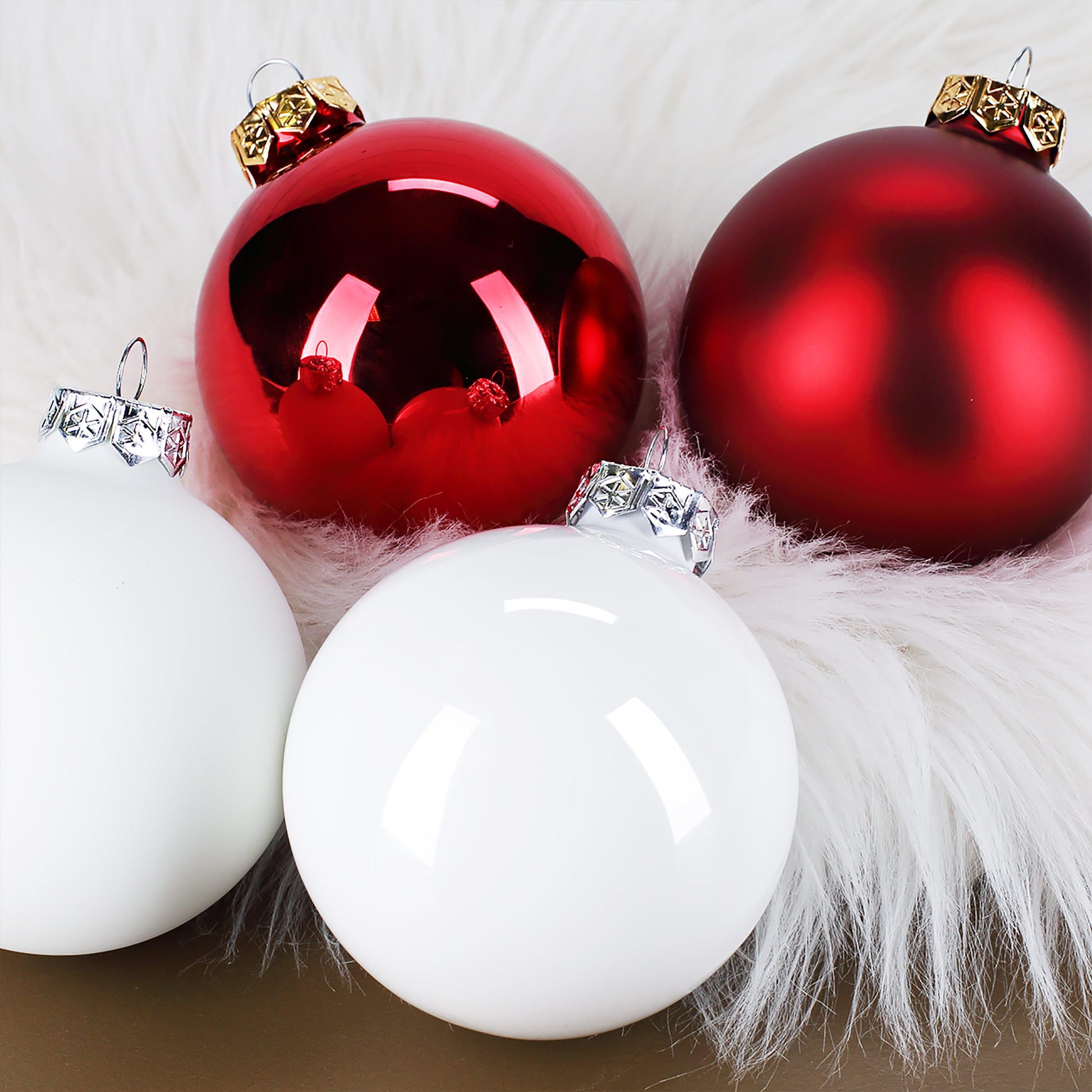 Vier Weihnachtsbaumkugeln in Rot und Weiß, matt und glänzend, liegend auf weißem Stoff, mit feiner Gravur und silberner Aufhängung.