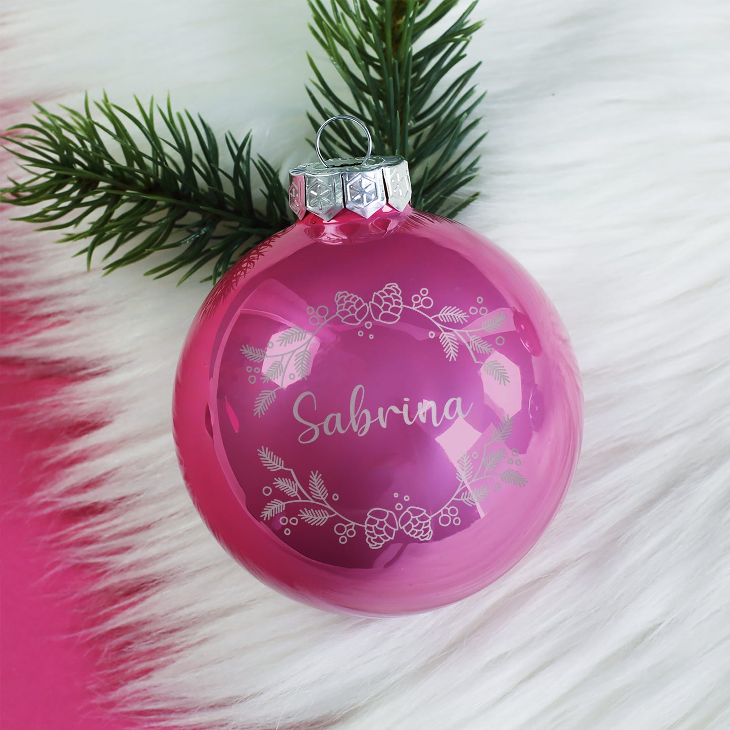 Rosa Weihnachtskugel mit Gravur „Sabrina“ und Kranzmotiv, glänzende Oberfläche, auf hellem Hintergrund arrangiert.