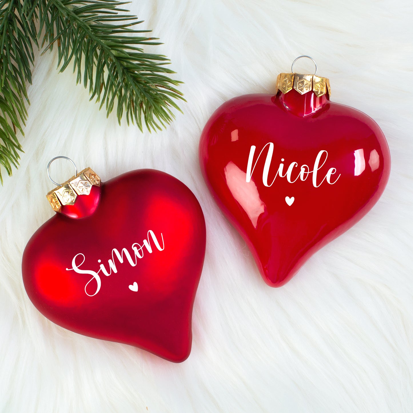 Zwei personalisierte Herz-Christbaumkugeln (matt „Simon“, glänzend „Nicole“) nebeneinander auf weißem Fell mit Tannenzweig.
