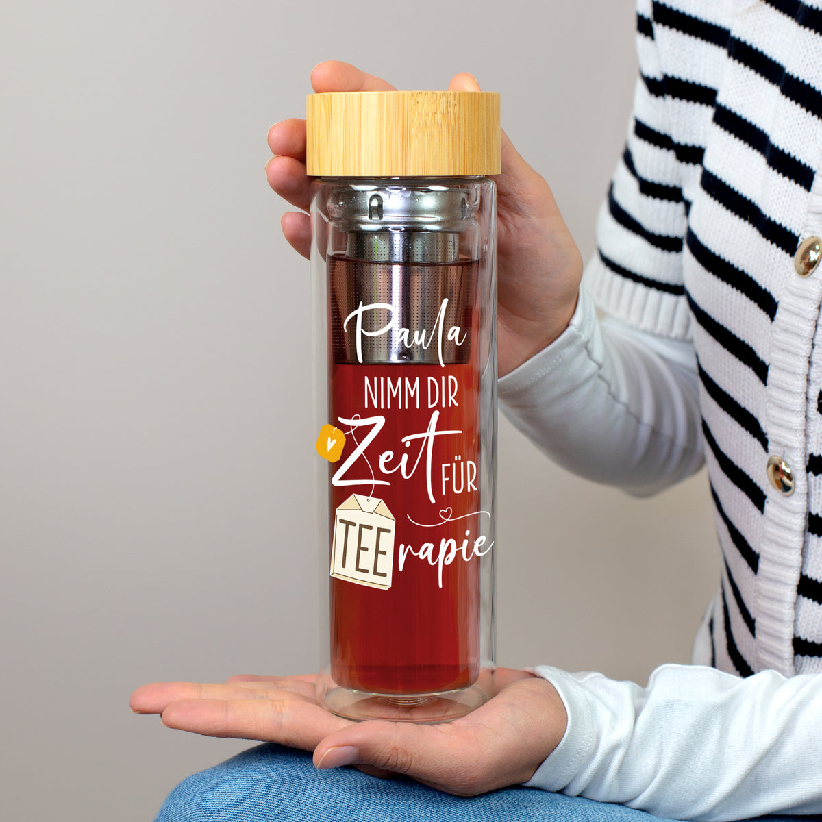 Personalisierte Teeflasche aus doppelwandigem Glas mit Bambusdeckel, Edelstahl-Teesieb und Aufdruck „Paula – Nimm dir Zeit für TEErapie“ in den Händen einer Person