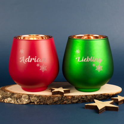 Zwei gravierte Windlichter in Rot und Grün mit Schneeflockenmotiv und Namen, auf Holzscheibe mit Sternen dekoriert.