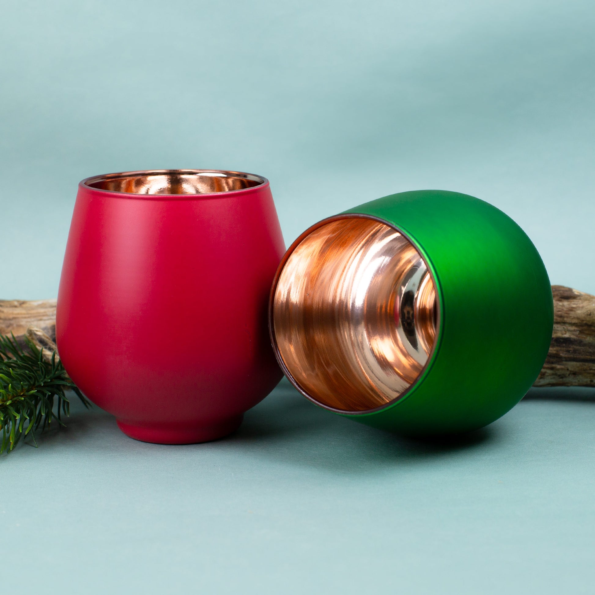 Rotes und grünes Windlicht mit graviertem Herzmotiv, dekorativ auf hellem Untergrund mit Holz und Tannenzweigen arrangiert.