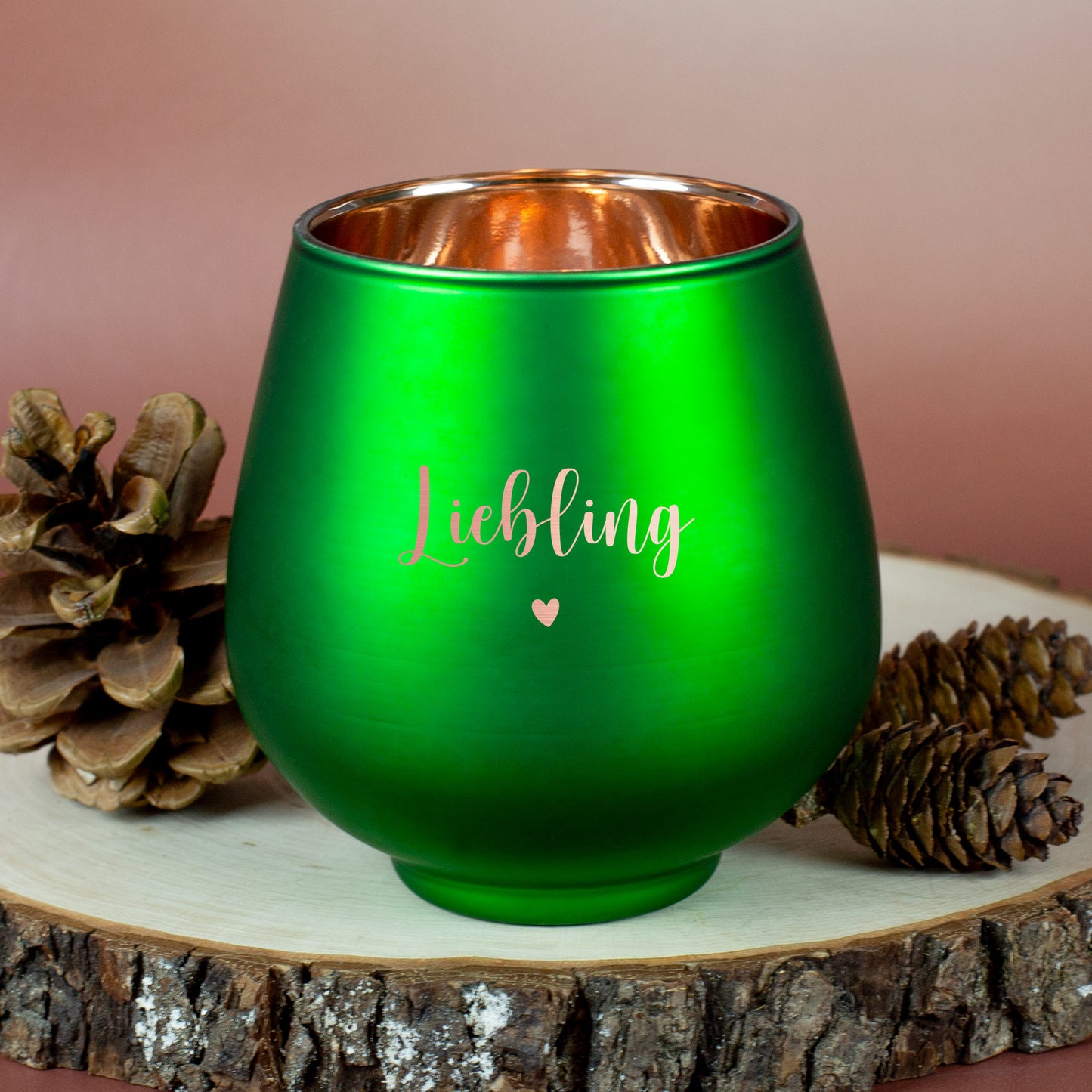 Grünes Windlicht mit Gravur „Liebling“ und Herzmotiv, auf Holzplatte mit Zapfen und winterlicher Deko platziert.