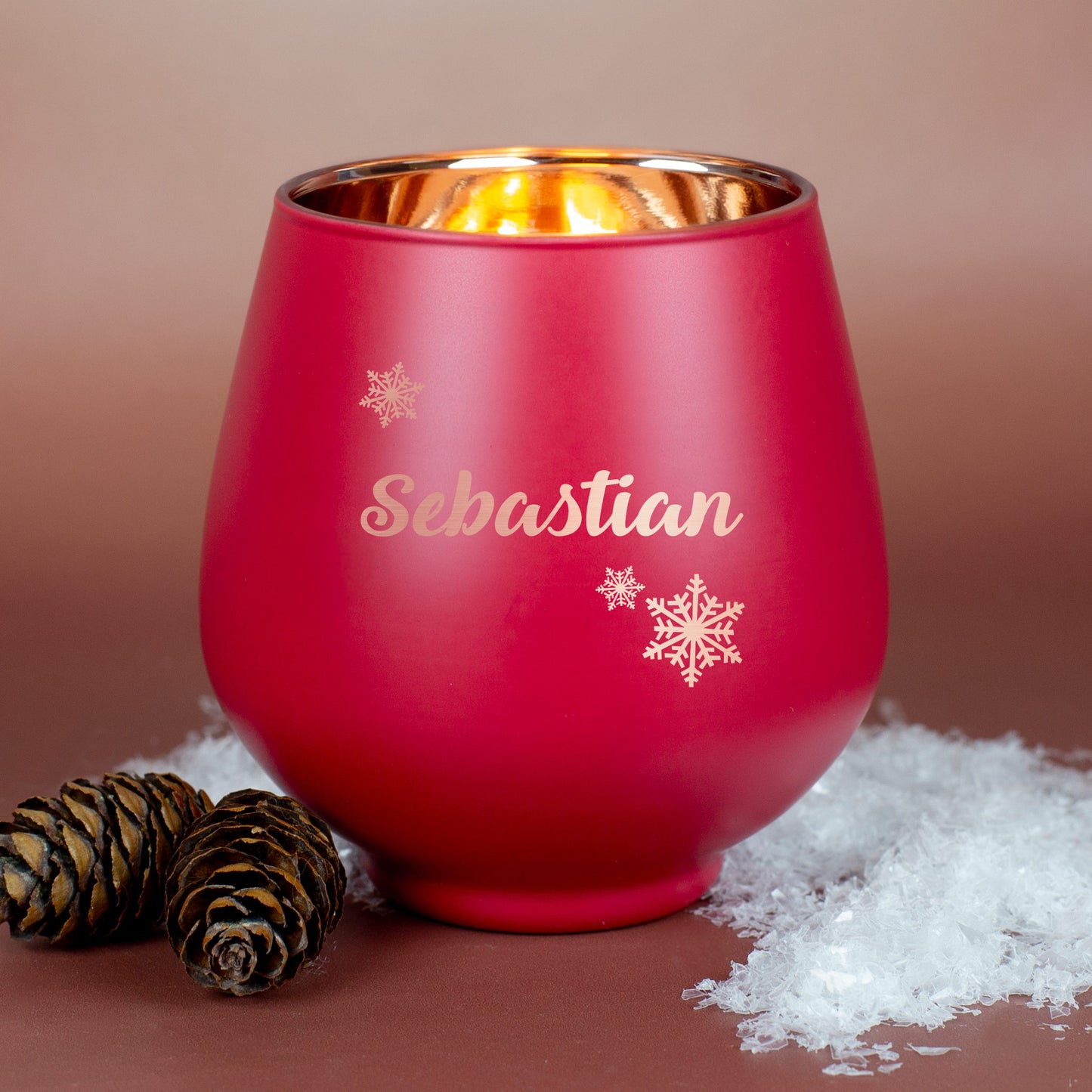 Rotes Windlicht mit Gravur „Sebastian“ und Schneeflockenmotiv, auf braunem Untergrund mit Kiefernzapfen und Kunstschnee dekoriert.