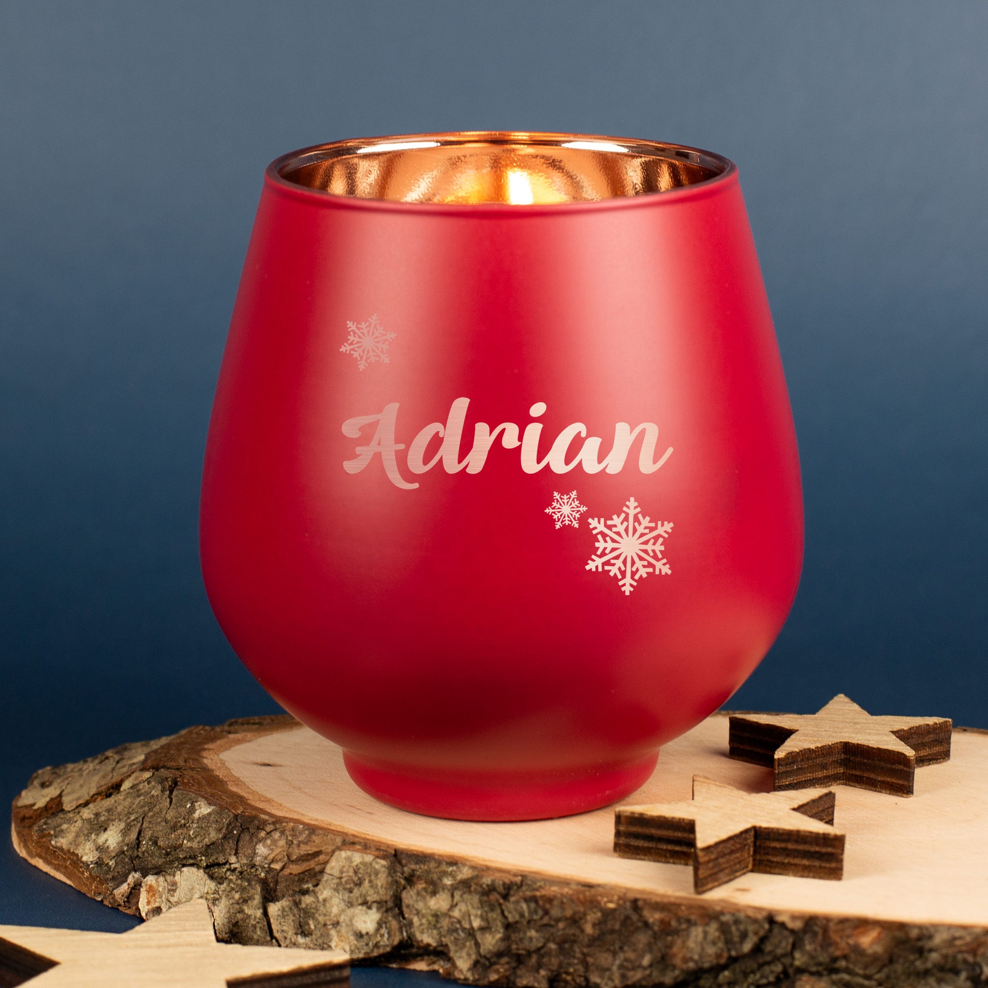 Rotes Windlicht mit Gravur „Adrian“ und Schneeflockenmotiv, auf Holzscheibe mit Sternen, im warmen Kerzenlicht leuchtend.