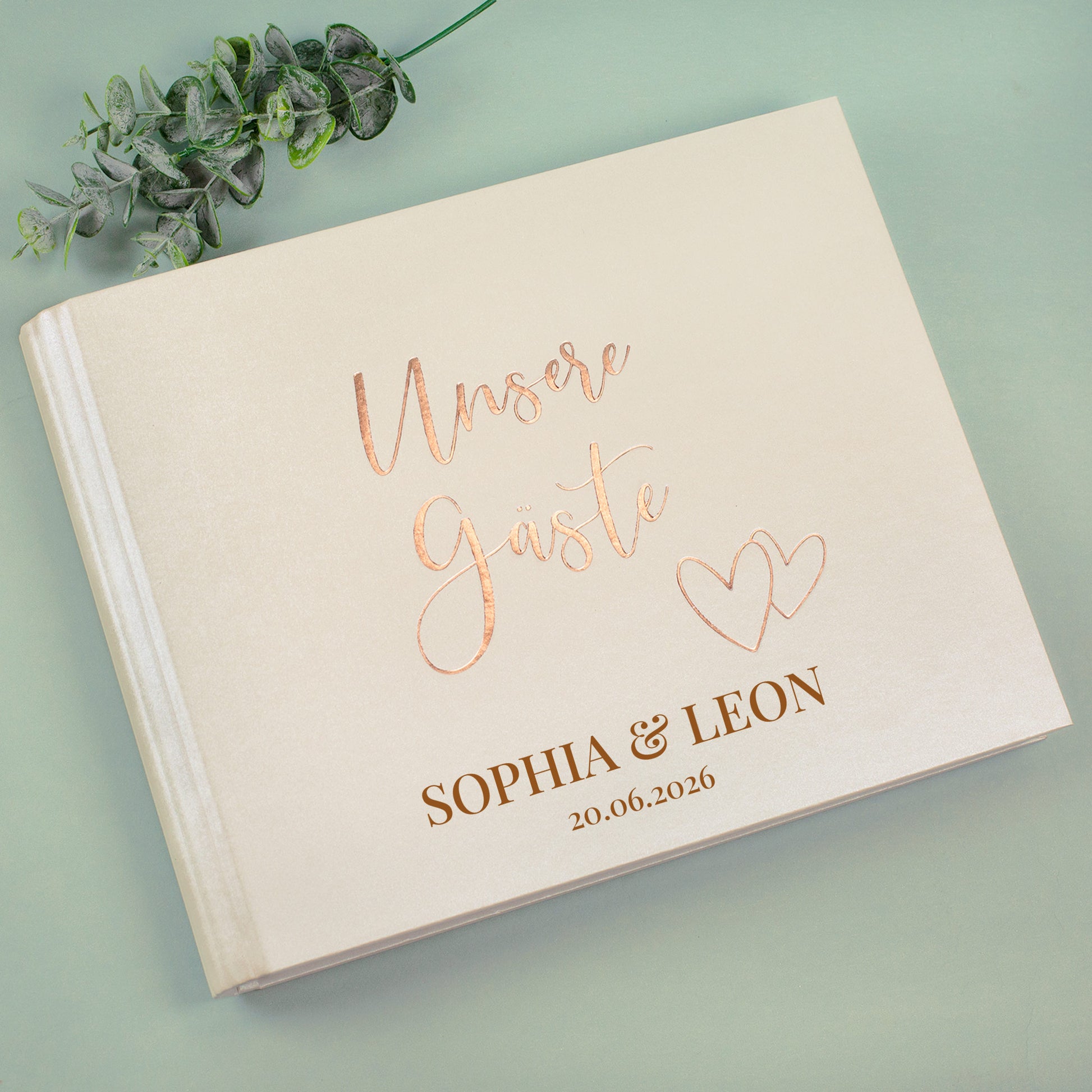 Cremefarbenes Gästebuch „Unsere Gäste“ mit Personalisierung, liegend mit Eukalyptus-Deko fotografiert.