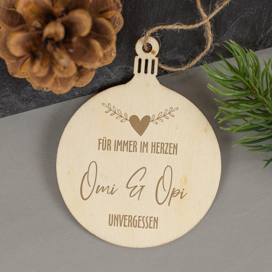 Gedenkanhänger aus Holz mit Gravur „Für immer im Herzen“, personalisiert mit Inschrift Omi & Opi, umgeben von Tannenzapfen und Zweigen.