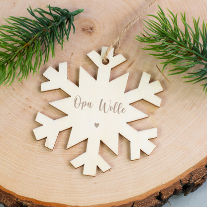 Holzanhänger Schneeflocke mit Gravur „Opa Wolle“ und Herzsymbol, liegend auf Holzscheibe mit Zweigen.
