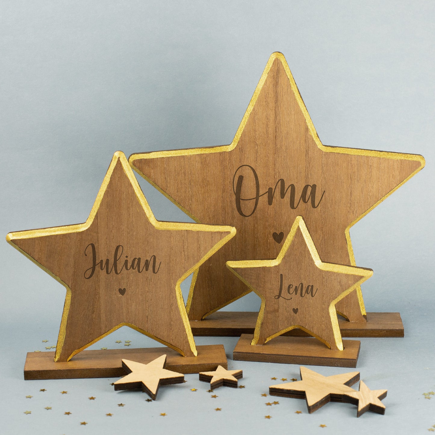 Drei Holzsterne mit goldenem Rand und Gravur der Namen „Oma“, „Julian“ und „Lena“, auf Holzsockeln arrangiert, umgeben von kleinen Sternstreuteilen.