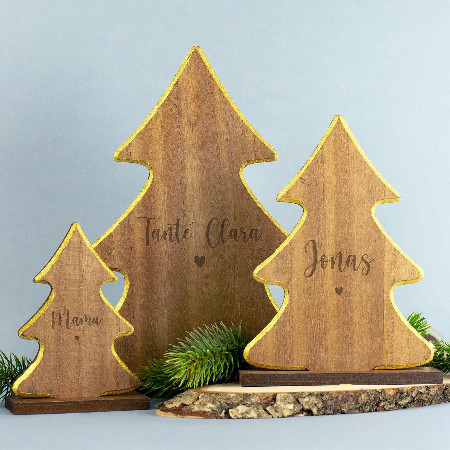 Drei Holzfiguren in Tannenbaumform mit goldener Kante, Gravuren „Mama“, „Tante Clara“ und „Jonas“, dekorativ auf Holzscheibe mit Tannengrün platziert.