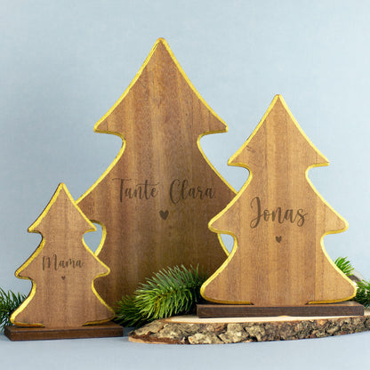 Drei Holzfiguren in Tannenbaumform mit goldener Kante, Gravuren „Mama“, „Tante Clara“ und „Jonas“, dekorativ auf Holzscheibe mit Tannengrün platziert.