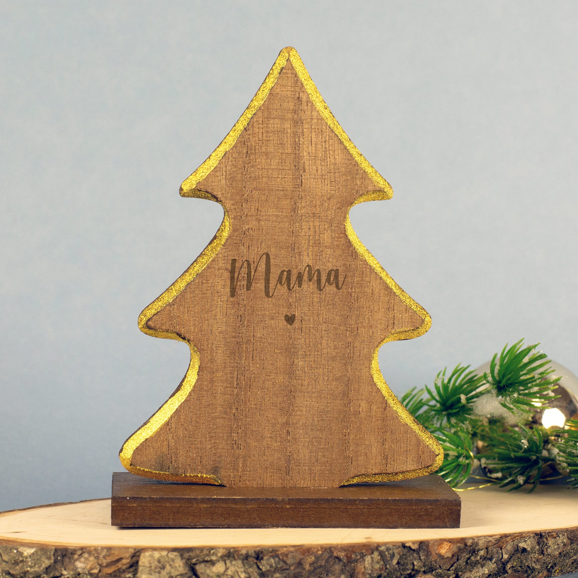 Einzelne Holzfigur in Tannenform mit Gravur „Mama“ und kleinem Herz, goldumrandet und auf einem Holzsockel stehend, im Hintergrund grüne Zweige und Lichter.
