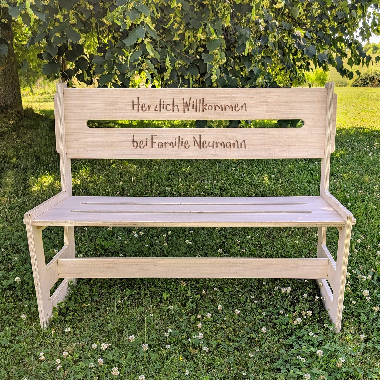 Holzbank mit Gravur „Herzlich Willkommen bei Familie Neumann“, aufgestellt im Garten auf grünem Rasen vor einem Baum
