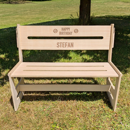 Frontalansicht einer gravierten Holzbank mit dem Namen 'Stefan' und den Zahlen 60 in Verkehrszeichenoptik – auf Rasen platziert
