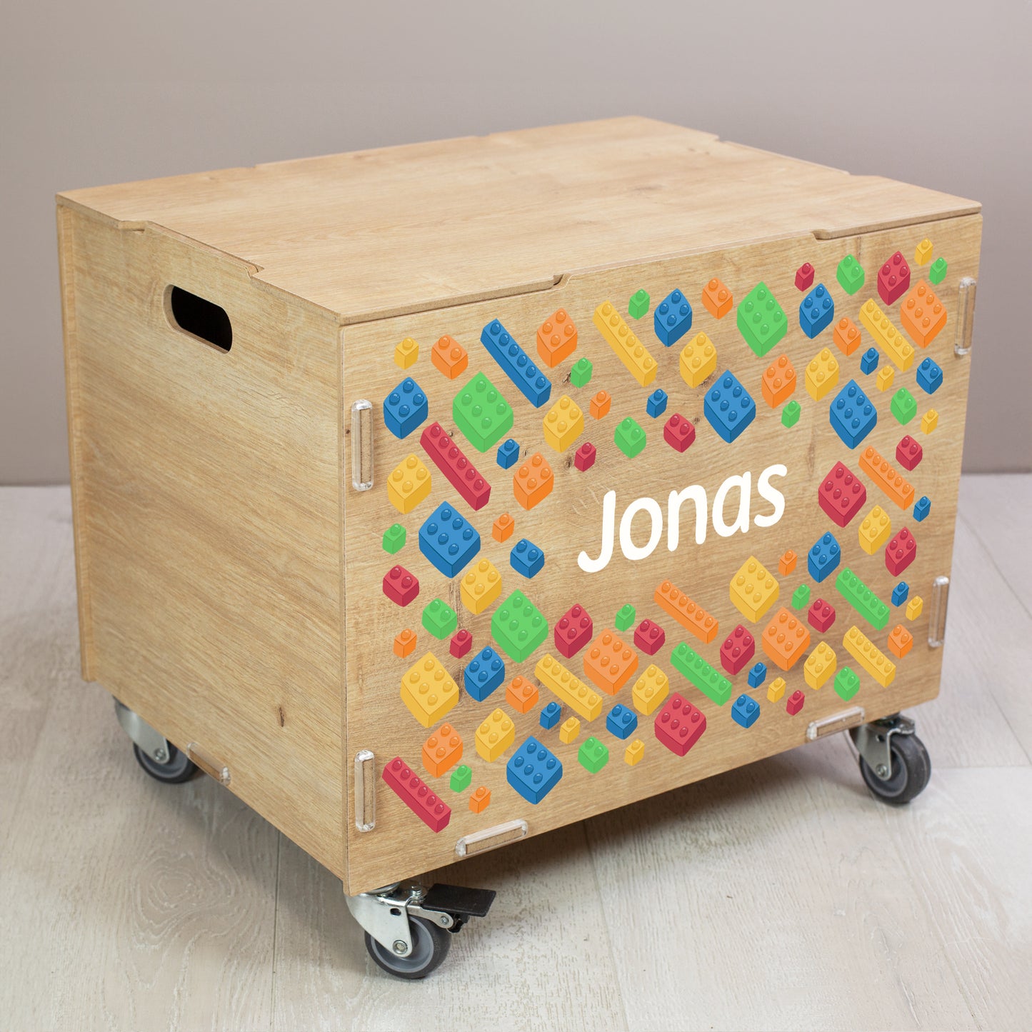 Personalisierte Rollbox aus Holz mit buntem Bausteinwelt-Motiv und Name, geschlossene Spielzeugkiste auf Rollen im Kinderzimmer