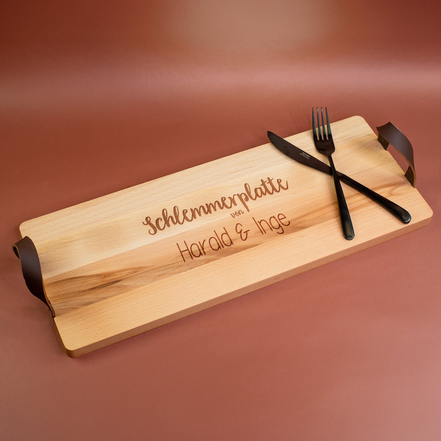 Personalisiertes Buchenholz-Servierbrett mit Gravur „Schlemmerplatte von Harald & Inge“ und seitlichen Ledergriffen, mit Besteck dekoriert.