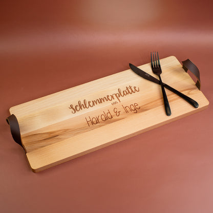 Personalisiertes Buchenholz-Servierbrett mit Gravur „Schlemmerplatte von Harald & Inge“ und seitlichen Ledergriffen, mit Besteck dekoriert.
