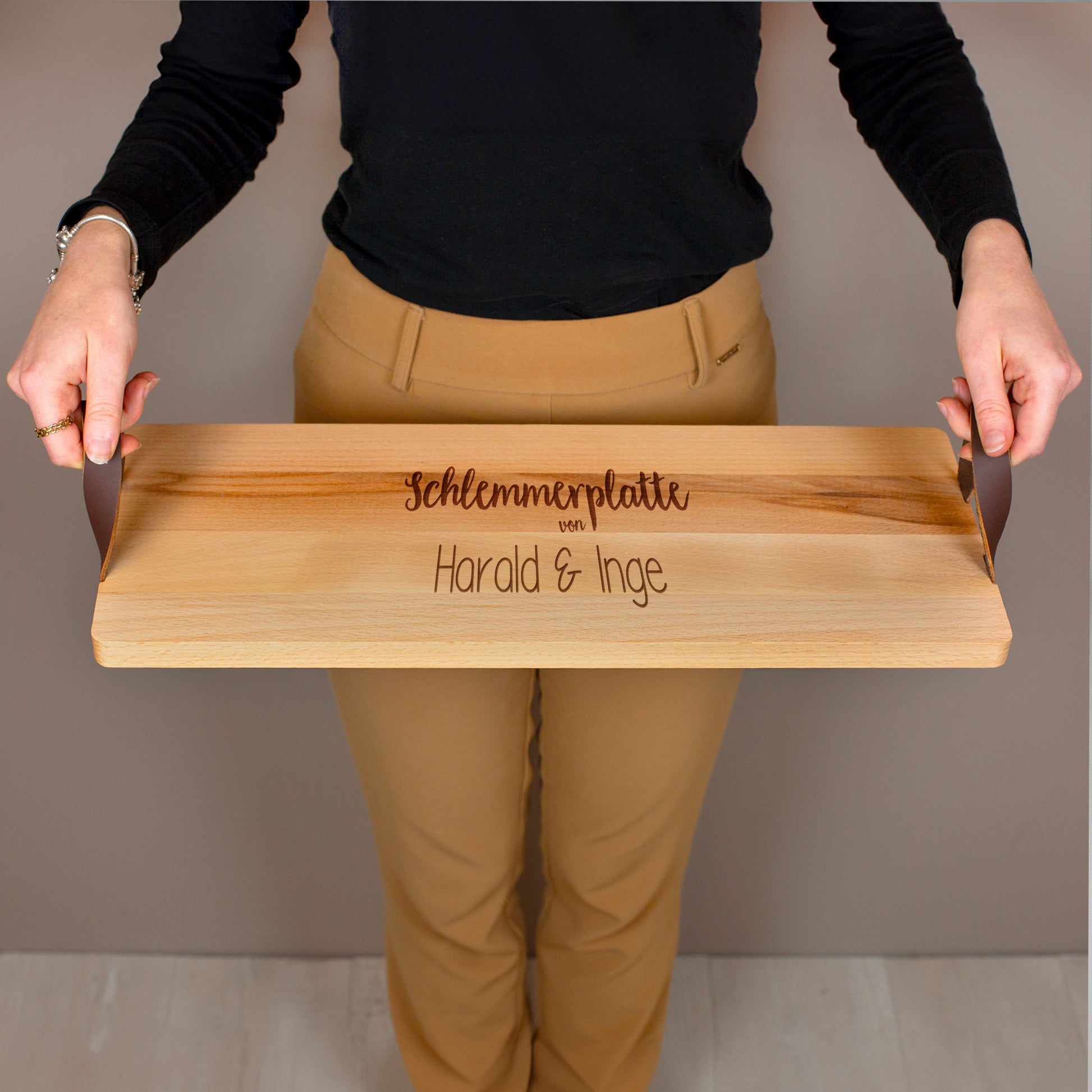 Servierbrett mit Ledergriffen im Einsatz, getragen von einer Person, Gravur „Schlemmerplatte von Harald & Inge“.