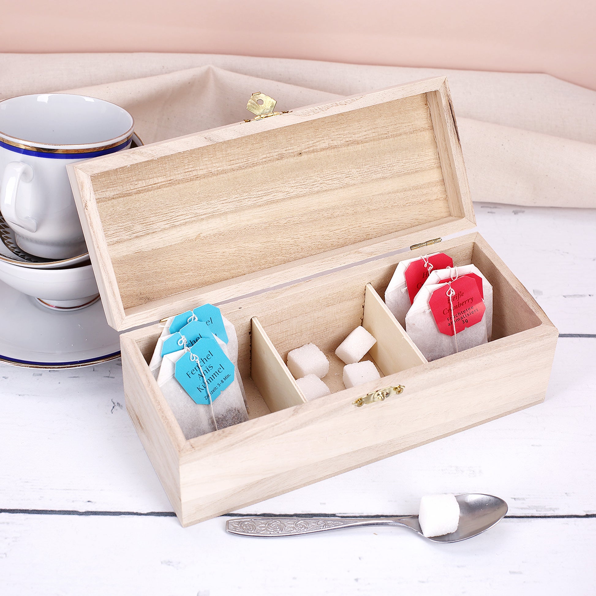 Offene Teebox aus Holz mit drei Fächern, befüllt mit Teebeuteln und Zuckerwürfeln auf hellem Tisch.