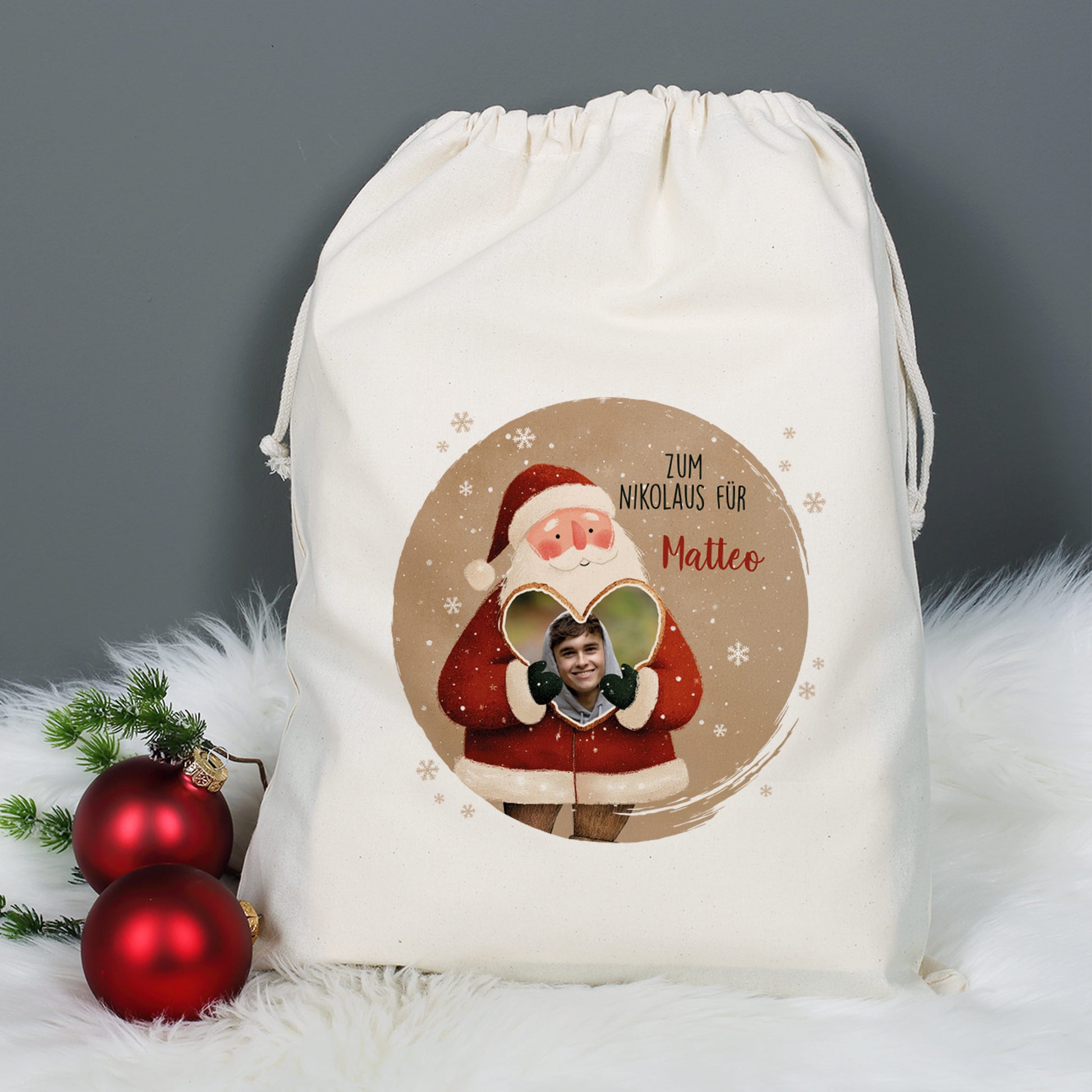 Weihnachtlicher Baumwollsack mit Weihnachtsmann-Motiv, Foto im Herz und Text „Zum Nikolaus für Matteo“, auf grauem Hintergrund mit weißen Fell-Details und Christbaumkugeln dekoriert.