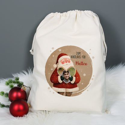 Weihnachtlicher Baumwollsack mit Weihnachtsmann-Motiv, Foto im Herz und Text „Zum Nikolaus für Matteo“, auf grauem Hintergrund mit weißen Fell-Details und Christbaumkugeln dekoriert.