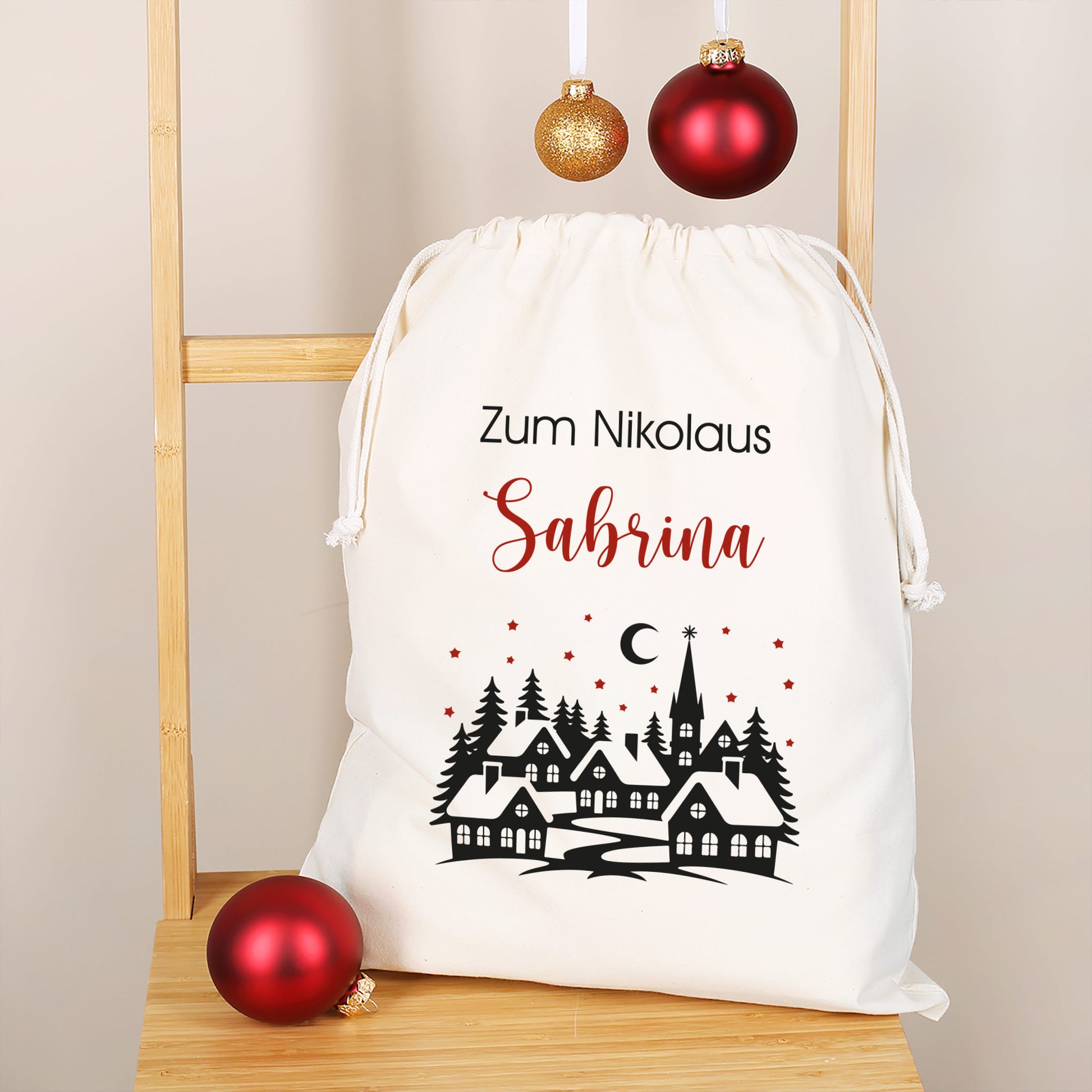 Mittlerer Nikolausbeutel mit Wunschtext „Zum Nikolaus Sabrina“, verschneite Dorfkulisse mit Tannen und Sternenhimmel, goldrote Kugeln als Deko.