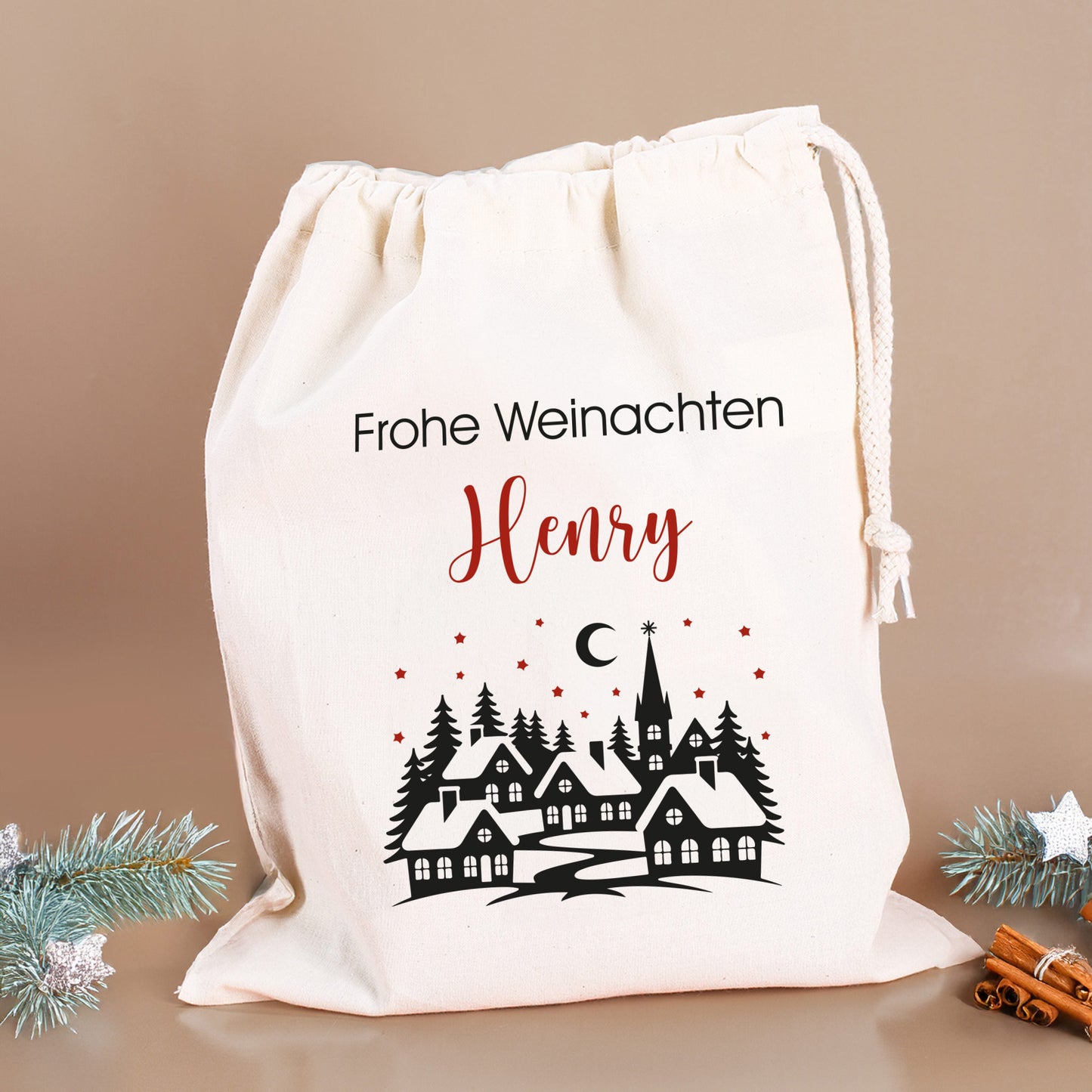 Mittlerer Geschenksack „Frohe Weihnachten Henry“ mit Winterdorf und Sternen, dekoriert mit Zimtstangen und silbernen Tannenzweigen.