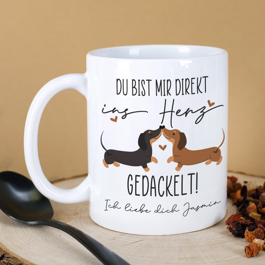 Personalisierte weiße Keramiktasse mit Dackel-Motiv und Spruch „Du bist mir direkt ins Herz gedackelt!“ auf Holzbrett, daneben Löffel und Tee