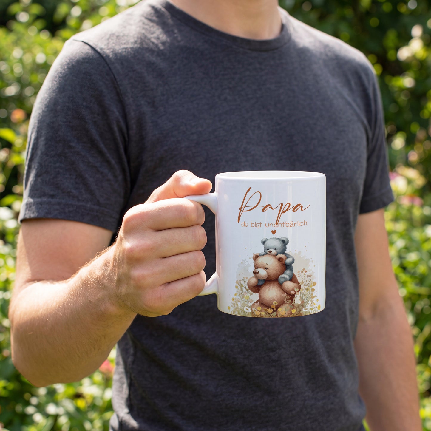 Ein Mann hält die weiße Tasse mit Bärenmotiv „Papa du bist unentbärlich“ in der Hand im Garten.
