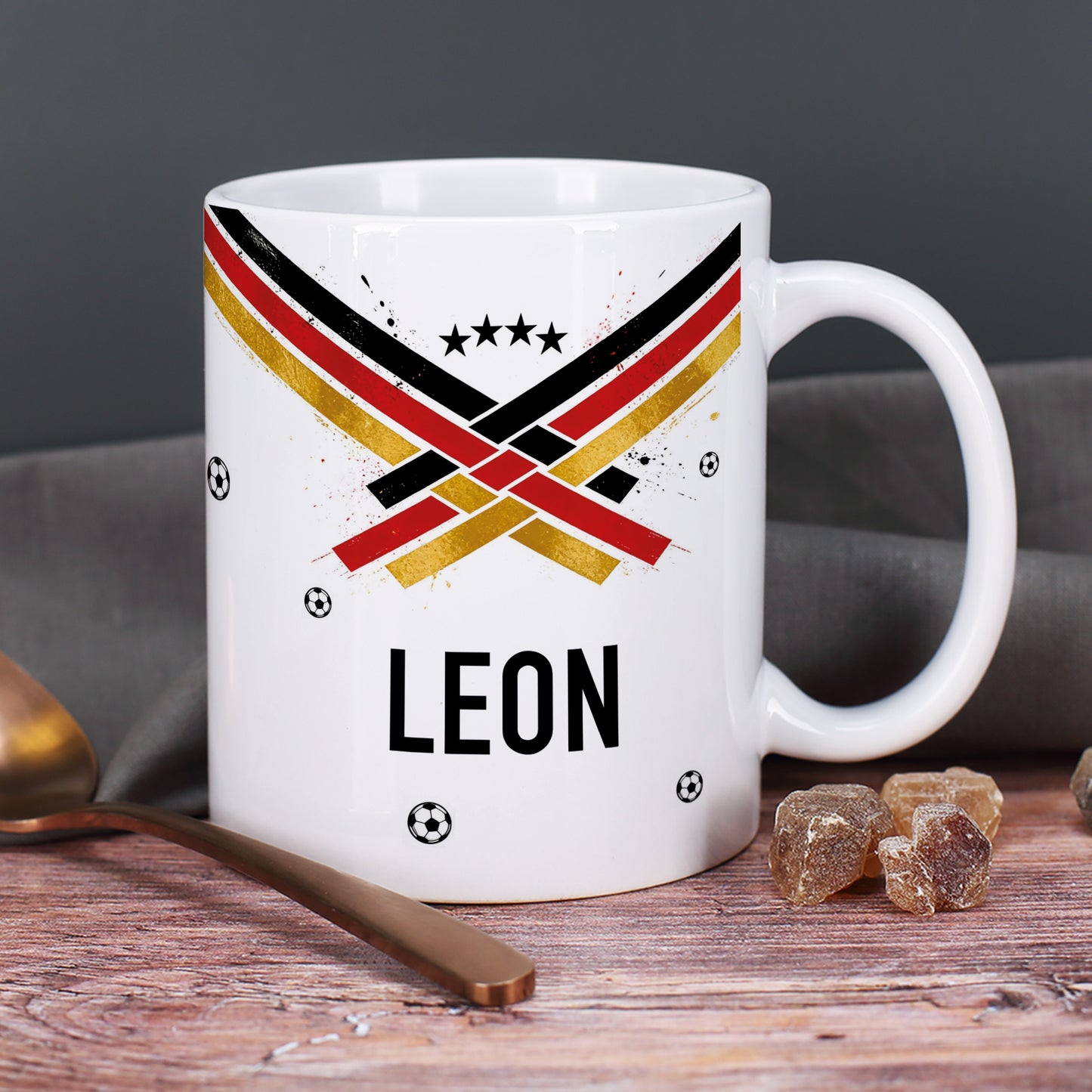 Personalisierte Fußball Tasse mit Name Leon, Fan-Design in Schwarz Rot Gold, kleine Fußbälle und Zuckerstücke im Hintergrund